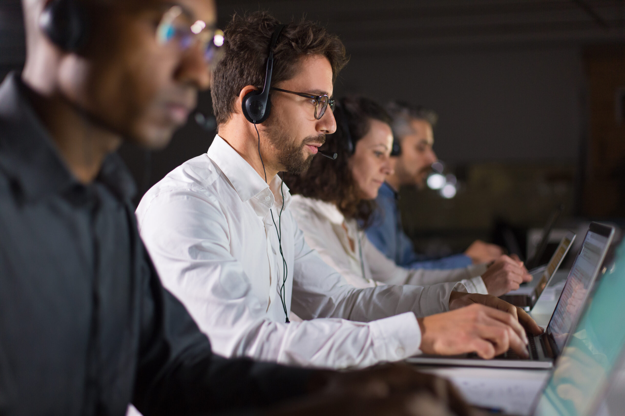 Side view of confident call center operator talking with client. Caucasian young man in eyeglasses typing on laptop while serving client. Call center concept